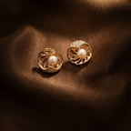 A pair of gold-toned stud earrings with a central white pearl and a halo of small clear stones, displayed on draped dark brown satin fabric.