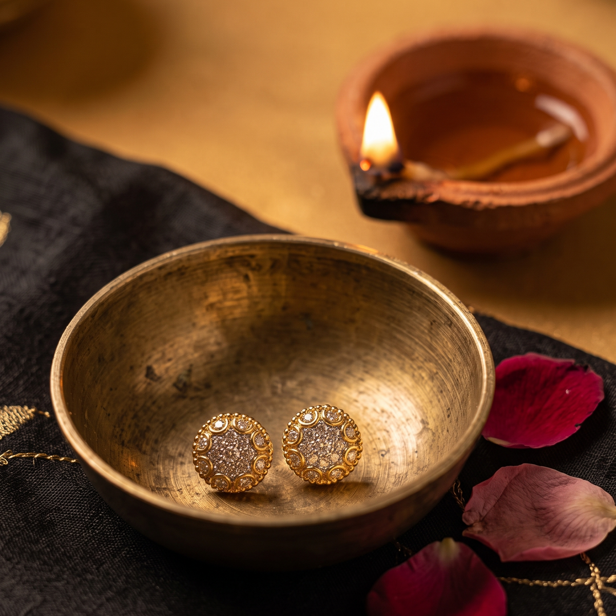 A pair of gold-toned stud earrings with clear stones, resting inside a small antique brass bowl. The bowl is placed on a black embroidered cloth with pink rose petals, with a lit clay diya in the warm background.