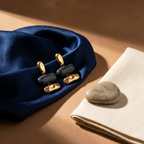 A pair of gold and dark blue drop earrings displayed on a navy blue satin fabric, styled with a beige linen napkin and a smooth stone on a warm brown background.