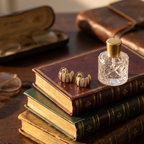 A pair of gold cufflinks with textured woven detailing rest on a stack of vintage leather-bound books next to a small glass perfume bottle. The scene is styled on a wooden table with antique eyeglasses in the background.
