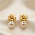 A pair of gold flower stud earrings with a white pearl drop, displayed on a draped beige satin fabric background.