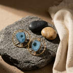 A pair of gold hoop earrings with blue agate stone inlays, styled on a textured gray stone with smooth river stones and a beige linen cloth.