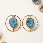 A pair of gold hoop earrings with irregular blue geode stone centers, displayed on a light cream background with decorative geode props.