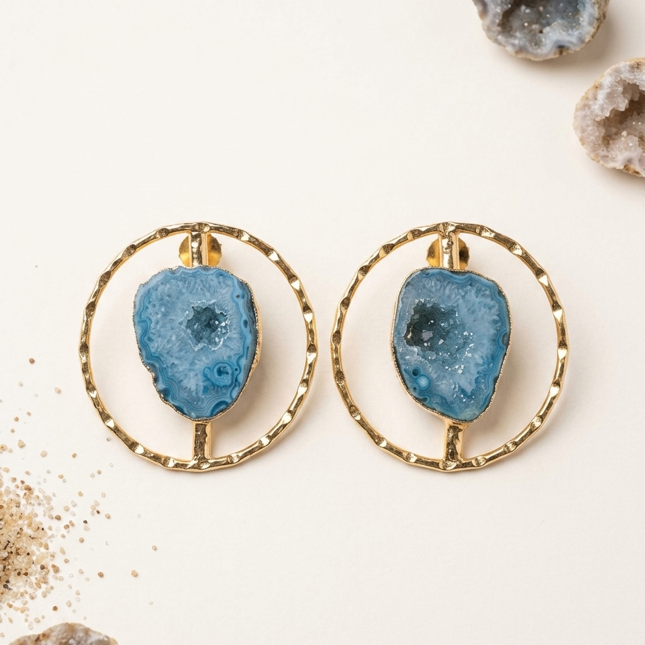A pair of gold hoop earrings with irregular blue geode stone centers, displayed on a light cream background with decorative geode props.