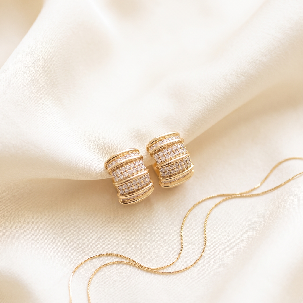 A pair of gold hoop earrings with rows of clear pavé stones, displayed on draped cream satin fabric with a matching gold snake chain.