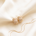 A pair of gold hoop earrings with rows of clear pavé stones, displayed on draped cream satin fabric with a matching gold snake chain.