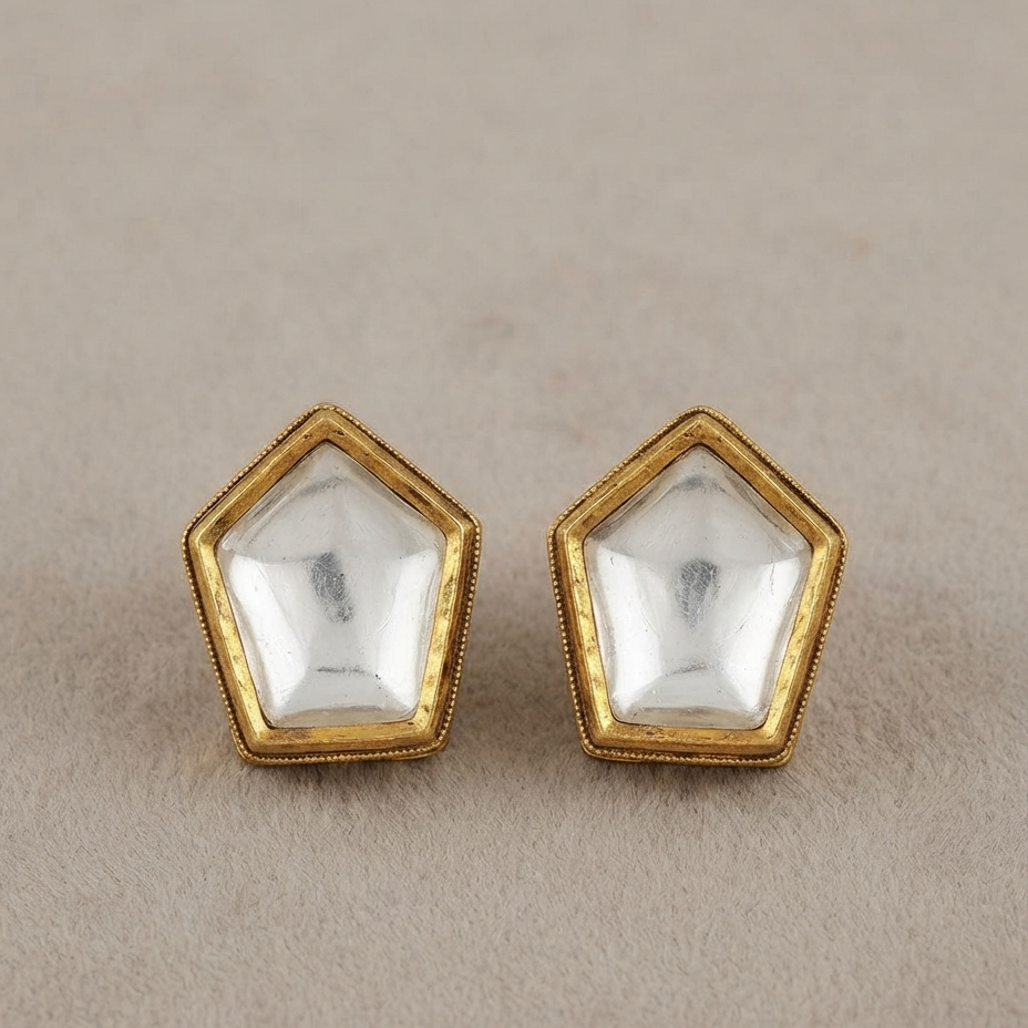 A pair of gold pentagonal stud earrings with clear faceted dome-cut stones, displayed on a textured beige surface.