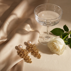 A pair of gold statement earrings with pearl-like round studs and cascading bead clusters, styled on draped champagne satin beside a clear coupe glass and a white rose.