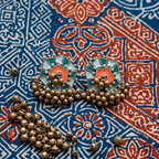 A pair of ornate Indian-style earrings with turquoise and coral enamel inlays, bronze-toned metal, and dangling bead chains, displayed on a blue and red patterned textile alongside loose beads.