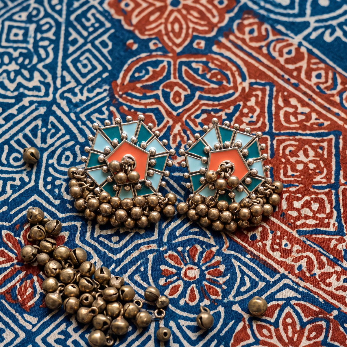 A pair of ornate Indian-style earrings with turquoise and coral enamel inlays, bronze-toned metal, and dangling bead chains, displayed on a blue and red patterned textile alongside loose beads.