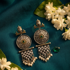 A pair of ornate antique-style silver earrings with circular filigree plates, small green stone centers, and dangling beaded chains, arranged on a deep teal fabric background with white flowers and green leaves.