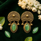 A pair of ornate antique bronze dangle earrings with green teardrop-shaped stones, styled on a dark green velvet background with white flowers and green leaves.