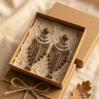 A pair of ornate dangling earrings in an open kraft gift box lined with textured paper, placed on top of a wrapped kraft box with twine, on a beige fabric surface with a dried leaf nearby.