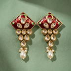 A pair of ornate gold-plated earrings with red enamel, green accents, clear stone details, and dangling white pearls, displayed on a textured sage green background.