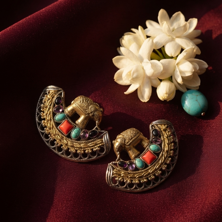 A pair of ornate gold-toned crescent-shaped earrings with turquoise, coral, and purple gemstone accents, displayed on deep burgundy fabric beside white flowers.