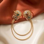 A pair of ornate gold-toned earrings with floral tops, multicolored enamel details, and long dangling chain loops, styled on draped satin fabric.