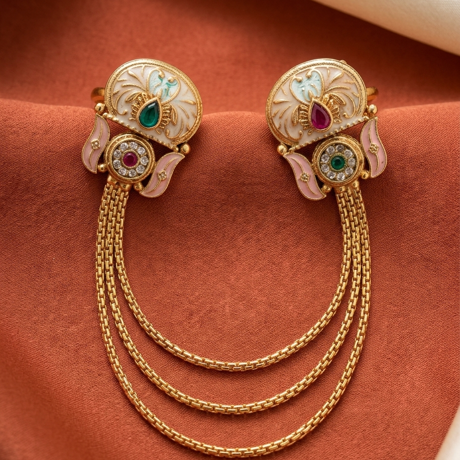 A pair of ornate gold-toned earrings with white enamel floral details, pink leaf accents, and colorful gemstone centers, connected by three draped chain strands, displayed on a textured rust-brown fabric background.