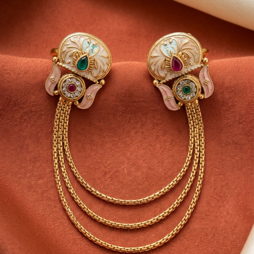 A pair of ornate gold-toned earrings with white enamel floral details, pink leaf accents, and colorful gemstone centers, connected by three draped chain strands, displayed on a textured rust-brown fabric background.