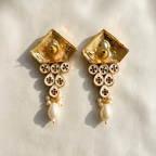 A pair of ornate gold-toned statement earrings with textured square tops, circular enamel details in red, green, and white, and dangling pearl-like beads, photographed on a light beige fabric background.