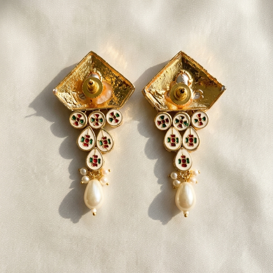 A pair of ornate gold-toned statement earrings with textured square tops, circular enamel details in red, green, and white, and dangling pearl-like beads, photographed on a light beige fabric background.