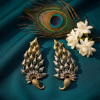A pair of ornate peacock-inspired earrings with gold and silver-toned metalwork, green stone accents, and small clear stones, arranged on a deep teal fabric backdrop with a peacock feather and white flowers.