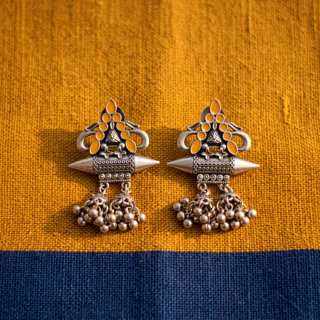 A pair of ornate silver-toned Indian jhumka earrings with yellow-gold inlay and dangling bead clusters, displayed on a textured fabric background split horizontally into mustard yellow on top and navy blue on the bottom.