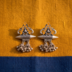 A pair of ornate silver-toned Indian jhumka earrings with yellow-gold inlay and dangling bead clusters, displayed on a textured fabric background split horizontally into mustard yellow on top and navy blue on the bottom.