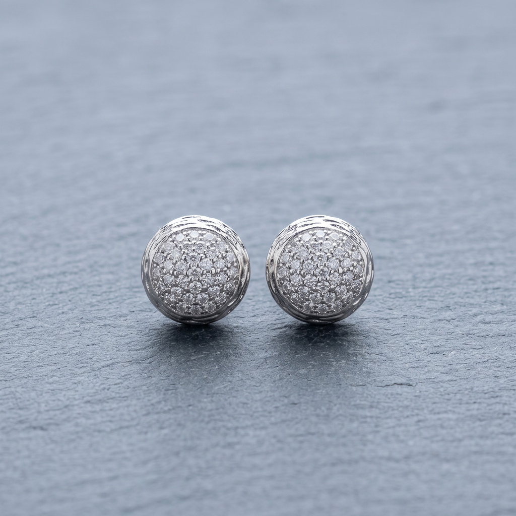 A pair of round silver stud earrings covered in small clear pavé stones, resting on a textured gray slate surface.