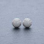 A pair of round silver stud earrings covered in small clear pavé stones, resting on a textured gray slate surface.