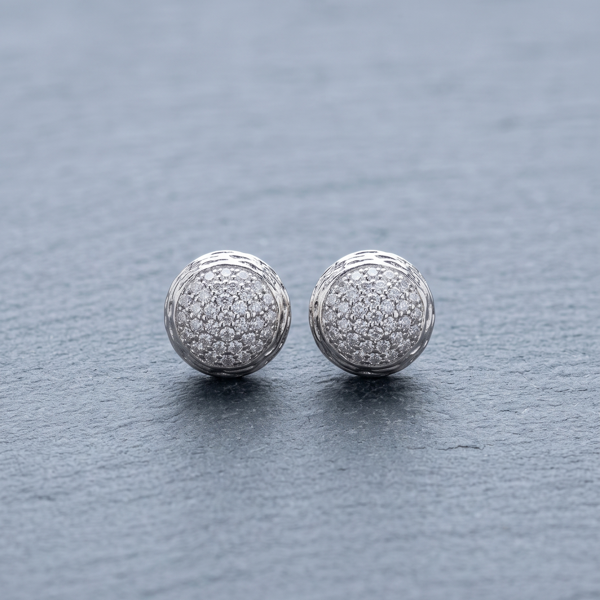 A pair of round silver stud earrings covered in small clear pavé stones, resting on a textured gray slate surface.