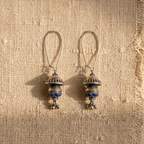 A pair of silver-toned dangle earrings with blue stone accents, displayed on a textured beige burlap fabric under warm sunlight.