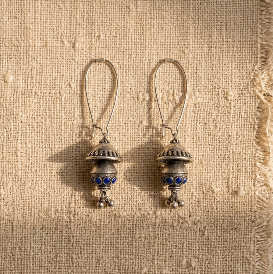 A pair of silver-toned dangle earrings with blue stone accents, displayed on a textured beige burlap fabric under warm sunlight.