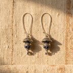 A pair of silver-toned dangle earrings with intricate filigree tops and dark blue enamel accents, resting on a textured beige linen fabric under warm sunlight.