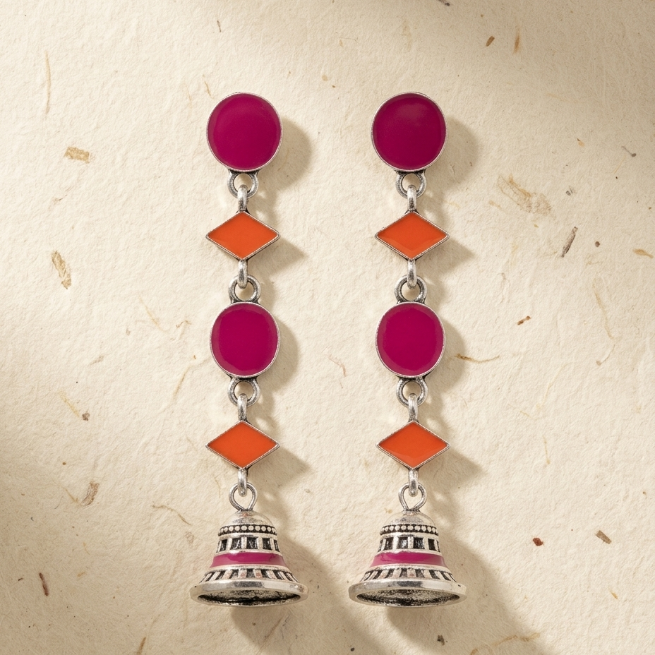 A pair of silver-toned dangle earrings with magenta pink circular stones, orange diamond-shaped stones, and bell-shaped dangling elements, displayed on a textured beige background.