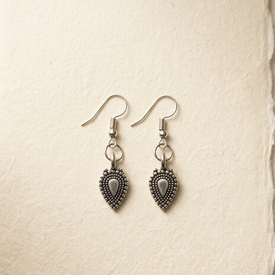 A pair of silver-toned dangle earrings with ornate, textured teardrop-shaped charms, displayed on a textured beige paper background.