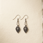 A pair of silver-toned dangle earrings with ornate, textured teardrop-shaped charms, displayed on a textured beige paper background.