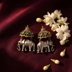 A pair of silver-toned elephant earrings with ornate gold-toned umbrellas and green stone accents, arranged on a deep burgundy satin fabric beside white flowers.