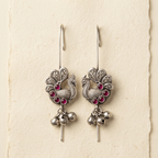 A pair of silver-toned peacock dangle earrings with small pink gemstone accents and dangling silver bells, displayed on a textured cream-colored card.