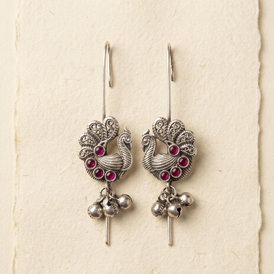 A pair of silver-toned peacock dangle earrings with small pink gemstone accents and dangling silver bells, displayed on a textured cream-colored card.