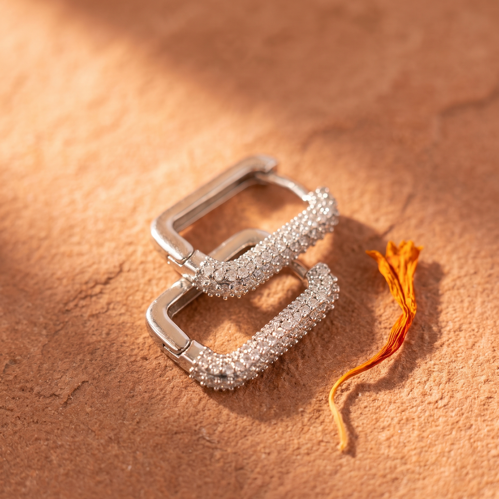A pair of silver-toned square hoop earrings covered in small clear stones, arranged on a warm brown textured surface with a small orange flower stem beside them.