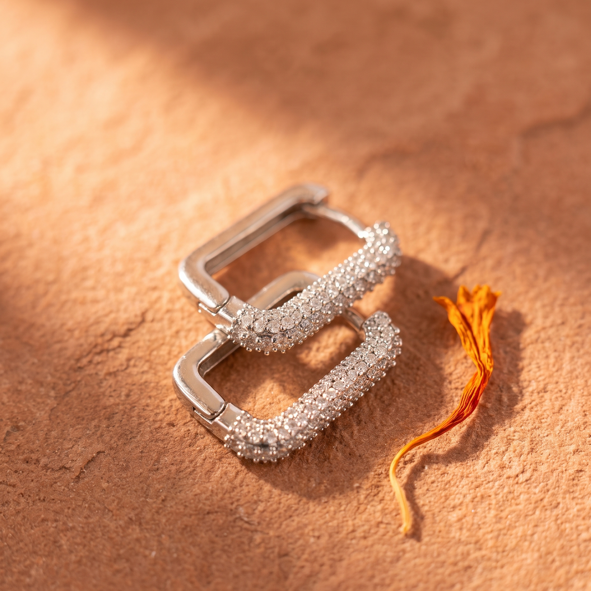 A pair of silver-toned square hoop earrings covered in small clear stones, arranged on a warm brown textured surface with a small orange flower stem beside them.