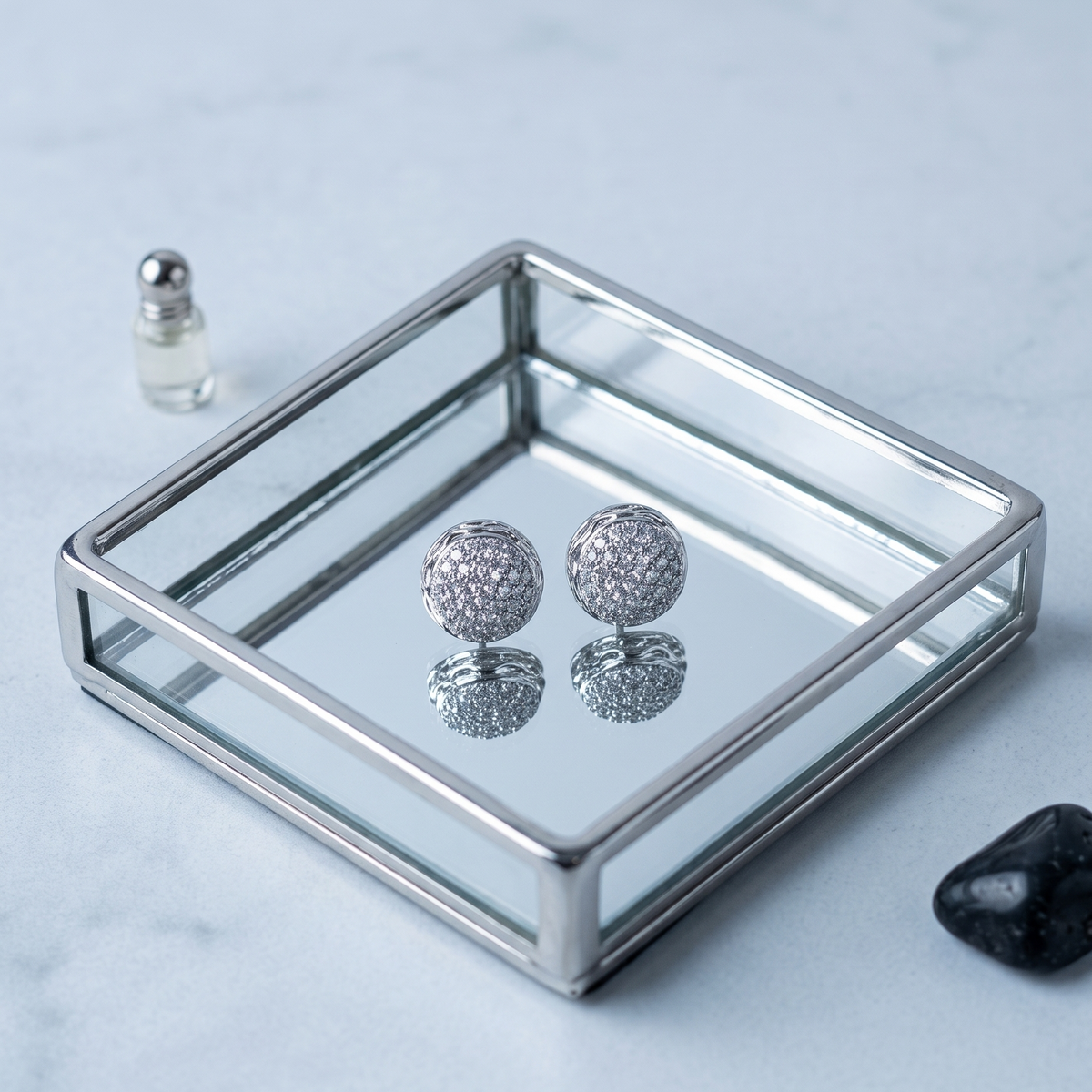 A pair of silver ball earrings with textured pavé-style detailing, displayed on a mirrored glass tray with a small perfume bottle and a black stone prop on a light marble surface.