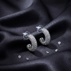 A pair of silver crescent-shaped hoop earrings covered in clear pavé stones, displayed on a draped black satin fabric with a few loose accent stones nearby.
