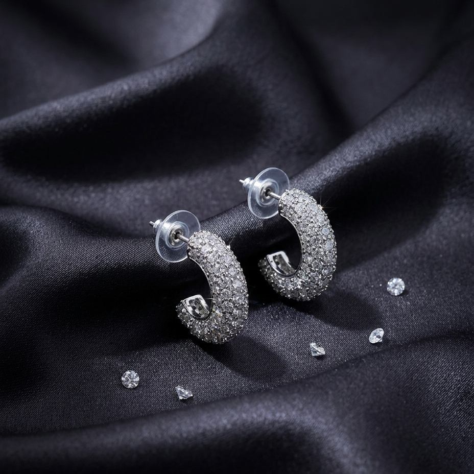 A pair of silver crescent-shaped hoop earrings covered in clear pavé stones, displayed on a draped black satin fabric with a few loose accent stones nearby.