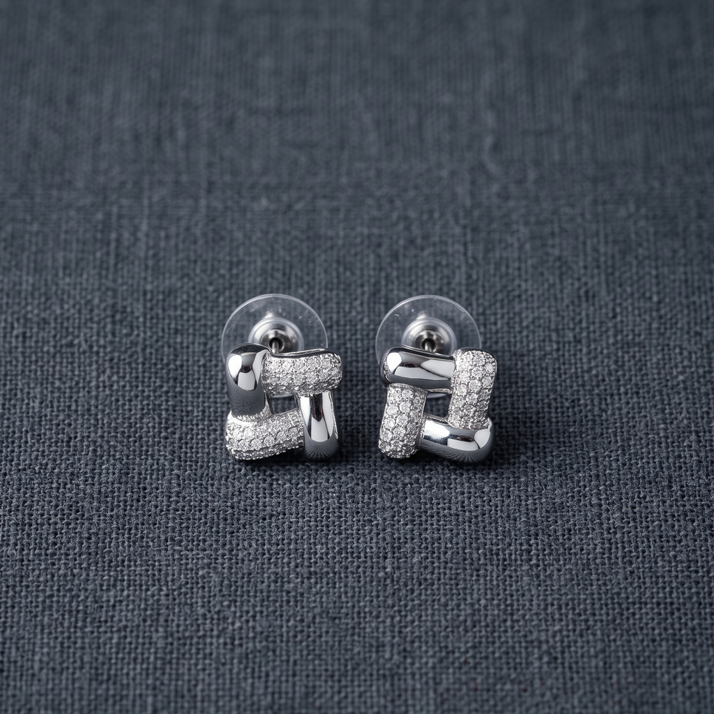 A pair of silver geometric stud earrings with pavé-set clear stones, displayed on a textured dark gray fabric background.