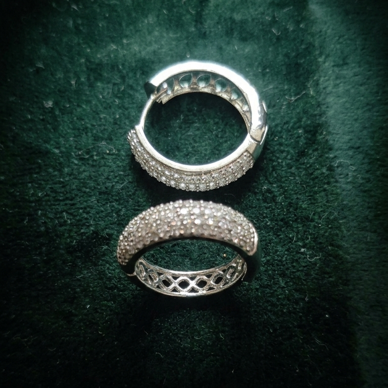 A pair of silver hoop earrings covered in small clear stones, with one earring showing the front and the other showing the back closure, placed on a dark green velvet background.
