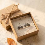 A pair of small bicycle-shaped stud earrings in dark metal, displayed inside an open kraft gift box lined with textured paper. A closed matching kraft box with twine sits behind it on a warm beige surface with a single dry leaf nearby.