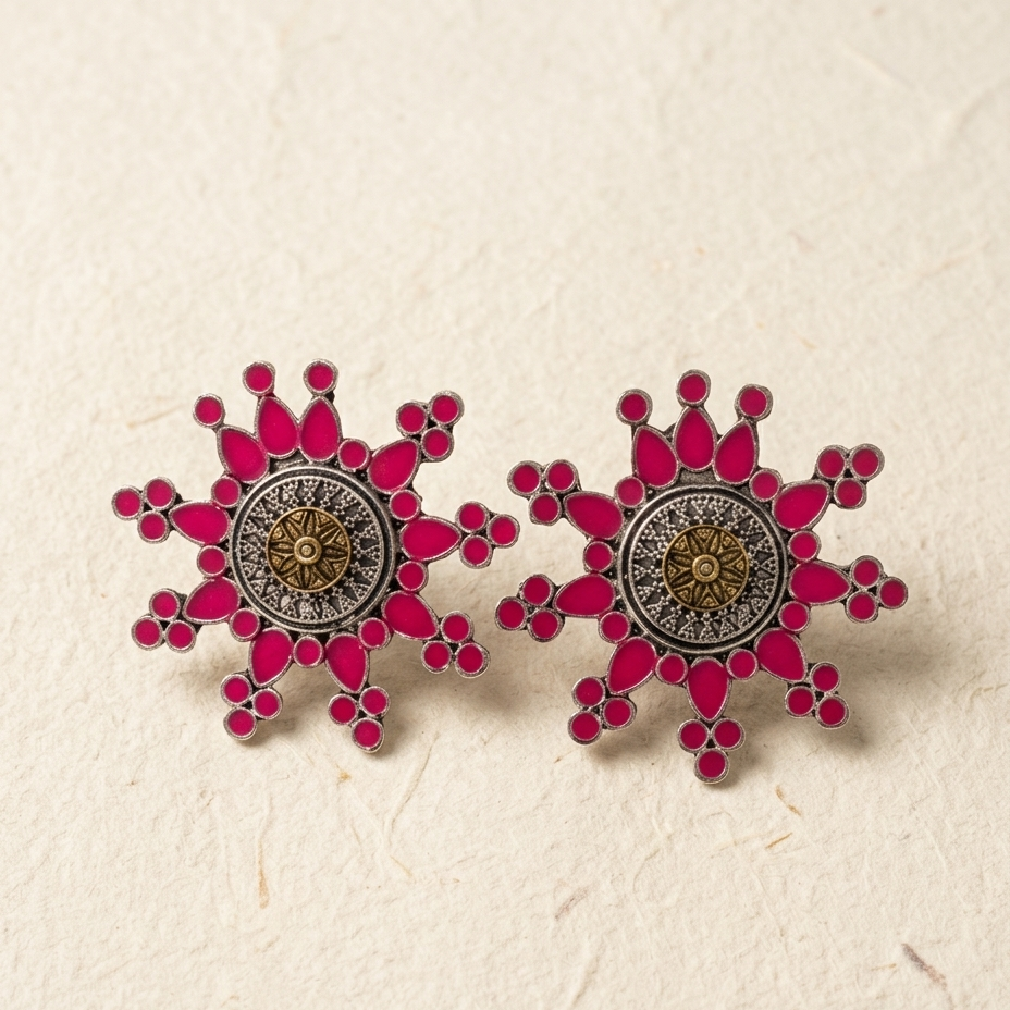 A pair of snowflake-shaped stud earrings with vibrant pink enamel, gold-toned central details, and silver-toned outer borders, displayed on a textured beige surface.