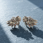 A pair of textured metallic leaf-shaped earrings with small clear stones, displayed on a slate surface with strong sunlight and shadows.