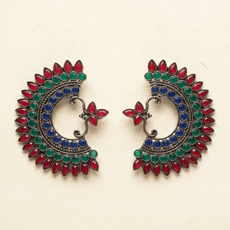 A pair of traditional Indian silver-toned earrings decorated with red, green, and blue stones, displayed on a textured off-white paper background.